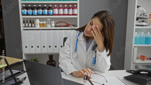 Wallpaper Mural Woman doctor sitting stressed in hospital clinic workplace with medical equipment in background appears exhausted with head resting on hand during workday. Torontodigital.ca