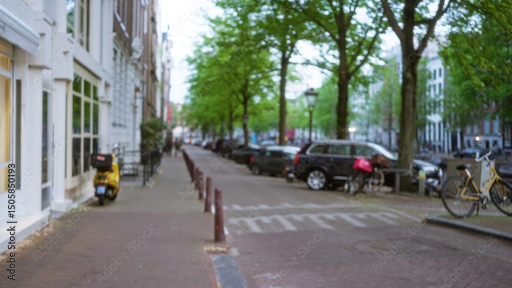 Obraz premium Blurry city street in amsterdam with bicycles and parked cars lined along the sidewalk under green trees, capturing the essence of urban life in the netherlands with a defocused view.