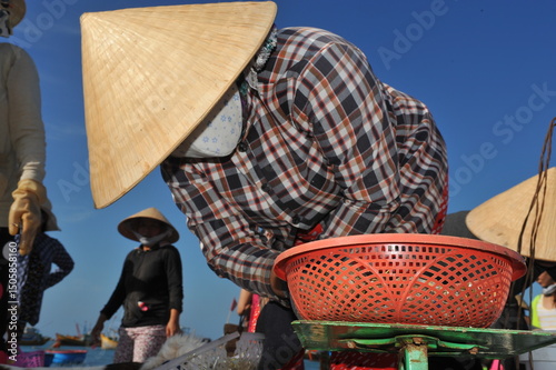 Unidentified fisher lady with conical hat looking at scale with fishing basket on top of it.
