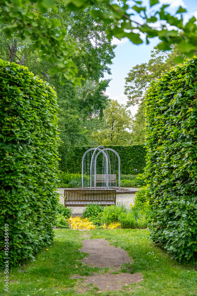 Obraz premium A picturesque garden path leads through tall, green hedges to a bench overlooking a modern water feature. The well-maintained grounds invite you to linger and convey a tranquil, natural atmosphere.