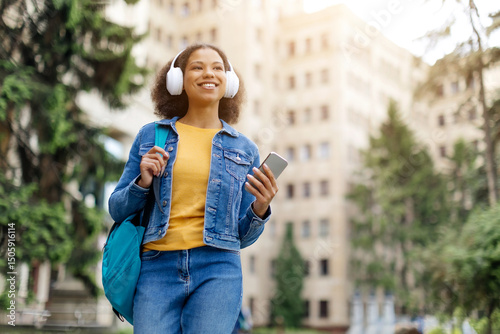 Joyful young African American student woman walking outdoors at campus with smartphone and headphones, happy black female listening favorite music, looking cheerful and focused, copy space