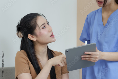 Asian female patient actively discussing dental care plan by pointing at digital tablet while speaking with dental assistant in blue scrubs during initial consultation at dental clinic.