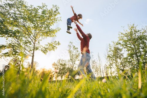 Foto Joyful father tossing young son into the air, playing in a sunny park, surrounded by trees and fresh green grass at sunset