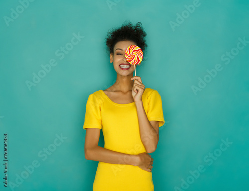 Foto Smiling curly mulatto girl in bright yellow dress holding candy lollipop