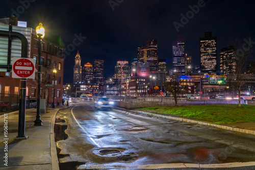 Night view of Boston skyline from a wet street with a car approaching.