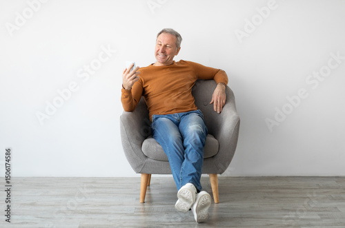 Quadro em tela Portrait of smiling mature man using mobile phone, watching video sitting resting on armchair isolated at white background wall