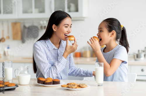 Dessert time for sweet tooth. Happy asian girl and young woman eating cookies and looking at each other, sitting at dinner table in kitchen, enjoying breakfast with milk