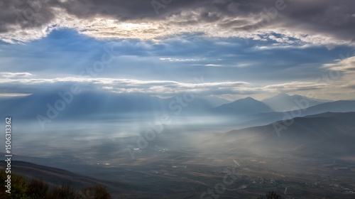 Serene sunlight piercing through dramatic clouds over a vast empty valley with layers of misty mountains in the distance, evoking peaceful landscape, wilderness, and natural atmosphere.
