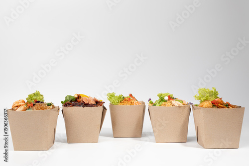 Five open eco-friendly brown paper boxes filled with various delicious and colorful Asian-style noodle and rice dishes, ready for takeaway , set against a clean white background.