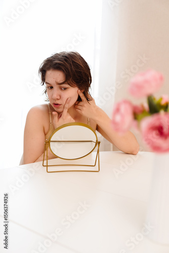 Stressed sad brunette young woman wearing beige top looking at mirror and touching her face having skin problems. Acne, pimples, wrinkles, dull skin