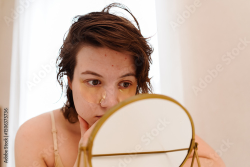 Stressed sad brunette young woman wearing beige top looking at mirror and touching her face having skin problems. Acne, pimples, wrinkles, dull skin