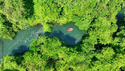 The aerial view captures the river as it weaves through the dense rainforest, with the raft drifting serenely, showcasing the peaceful coexistence of human activity and nature in a tropical paradise.
