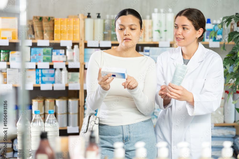 Obraz premium Slightly worried Asian woman in pharmacy checking medicine box while polite friendly young female pharmacist suggesting suitable ointment as treatment