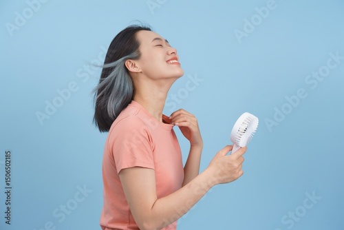 Woman enjoying a cool breeze while holding a portable fan against a light blue background during a warm sunny day