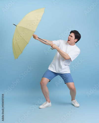 Young man playfully struggles with a yellow umbrella on a blue background during a sunny day