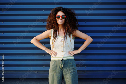 confident woman poses against blue wall wearing sunglasses and casual attire. emphasizes modern style and individuality. reflects urban fashion and carefree lifestyle in vibrant setting photo