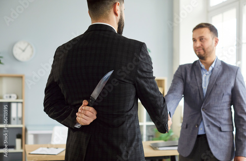 Businessman holding knife behind his back and shaking hands with male partner wearing suit standing in office. Man doing dishonest agreement. The concept of business betrayal and backstabbing.