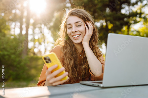 Wallpaper Mural Young woman with laptop and phone at wooden table in sunny park. Female freelancer working outdoors and texting on phone. Freelancing, nature concept. Torontodigital.ca