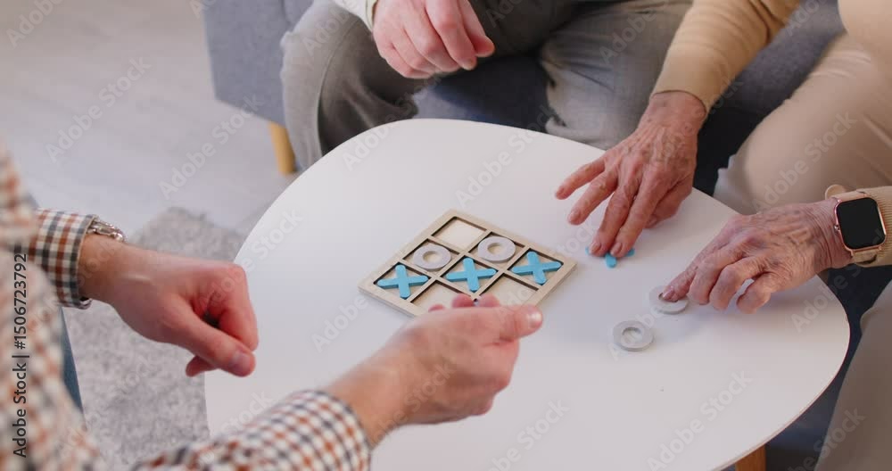 Social interaction. Three adults are enjoying game of tic-tac-toe, displaying their playful competitiveness. Millennial man and two mature people sit at small table with wooden tic-tac-toe board on it