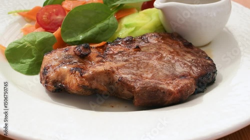 Juicy grilled pork steak served with fresh spinach leaves, carrot, cucumber, and cherry tomato salad on white plate in sunlight.
