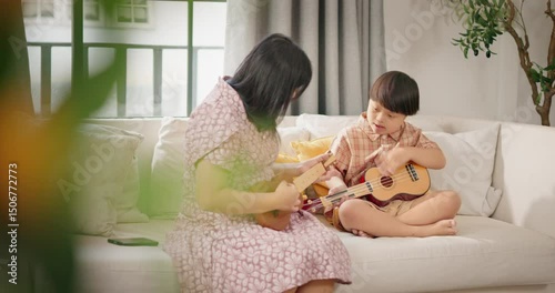 Asian young boy with Down syndrome playing ukulele guitar with his mother on a soft white sofa in a warm home. Family bonding, inclusive parenting, childhood creativity, and music therapy.