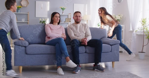 Exhausted and tired parents sitting on the couch while kids playing, run around, and create noise at home. Misbehave, chaos, stress, and challenges of parenting, balanced with moments of family life.