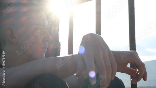 A shaved-head teenage boy sits quietly by a barred prison window, staring out with a sad, thoughtful gaze. A young inmate reflecting on his past, regretful and hoping for a second chance.