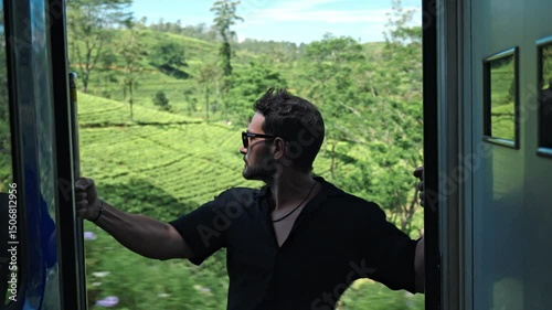 A man sits on the open door of a train traveling the scenic Ella-Kandy route in Sri Lanka. Behind him, expansive tea plantations spread across rolling hills, showcasing the region’s iconic landscape.