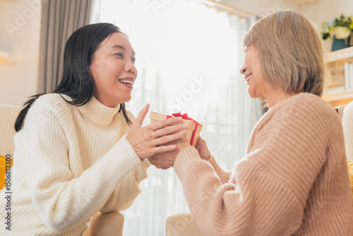 A loving asian daughter gives her mother a birthday gift while sitting together on the couch, exchanging a present box with warm smiles and a heartfelt embrace a perfect Mother's Day family moment.