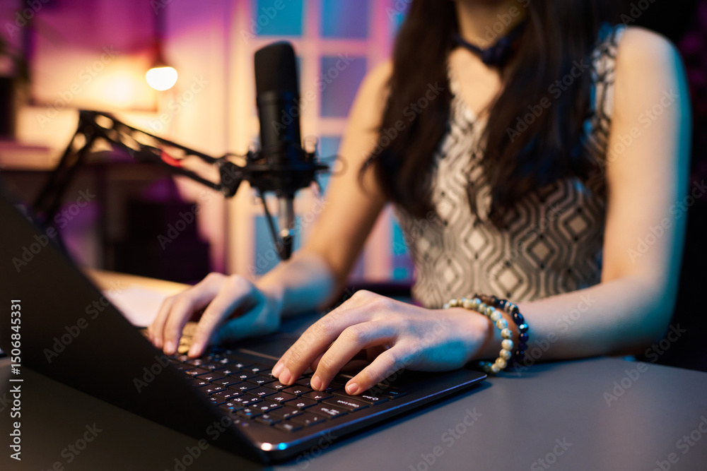 Fototapeta premium Young woman working on laptop with microphone in modern home studio with colorful lighting, focusing on task. Led lighting providing vibrant illumination enhancing workspace ambiance