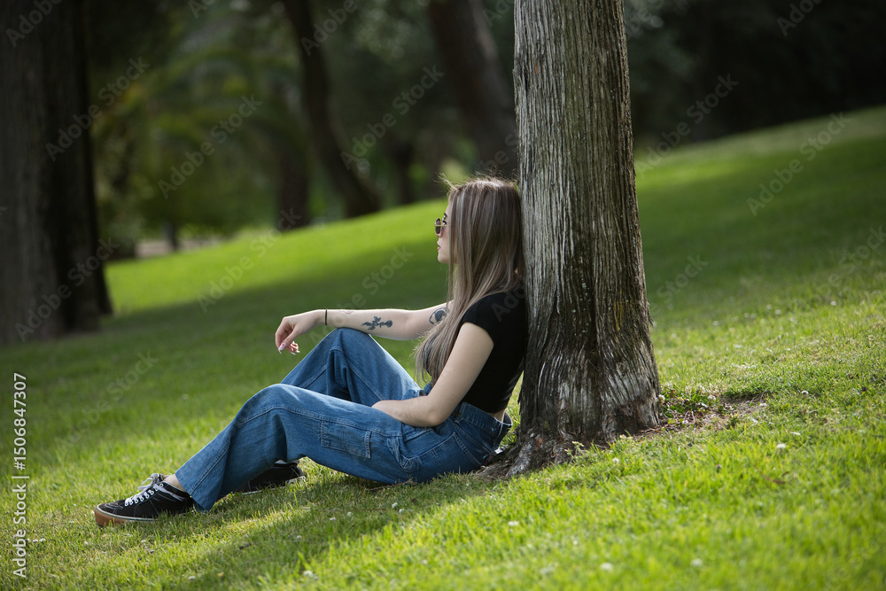 Obraz premium Young, beautiful blonde woman, wearing a black t-shirt, jeans, and sunglasses, relaxed, calm, and solitary, sitting on the grass leaning against a tree resting in a park. Concept of solitude, peace.