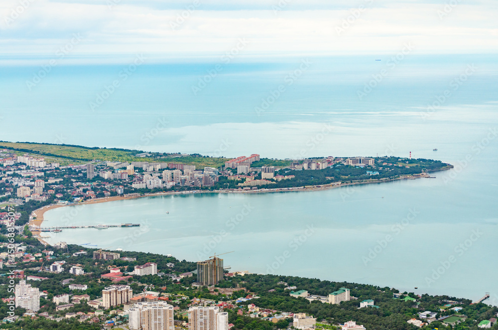 Fototapeta premium Aerial view showcasing Gelendzhik, russia, with coastal line curving along blue sea. City buildings and lush greenery highlight vibrant landscape. Soft lighting enhances scenic beauty