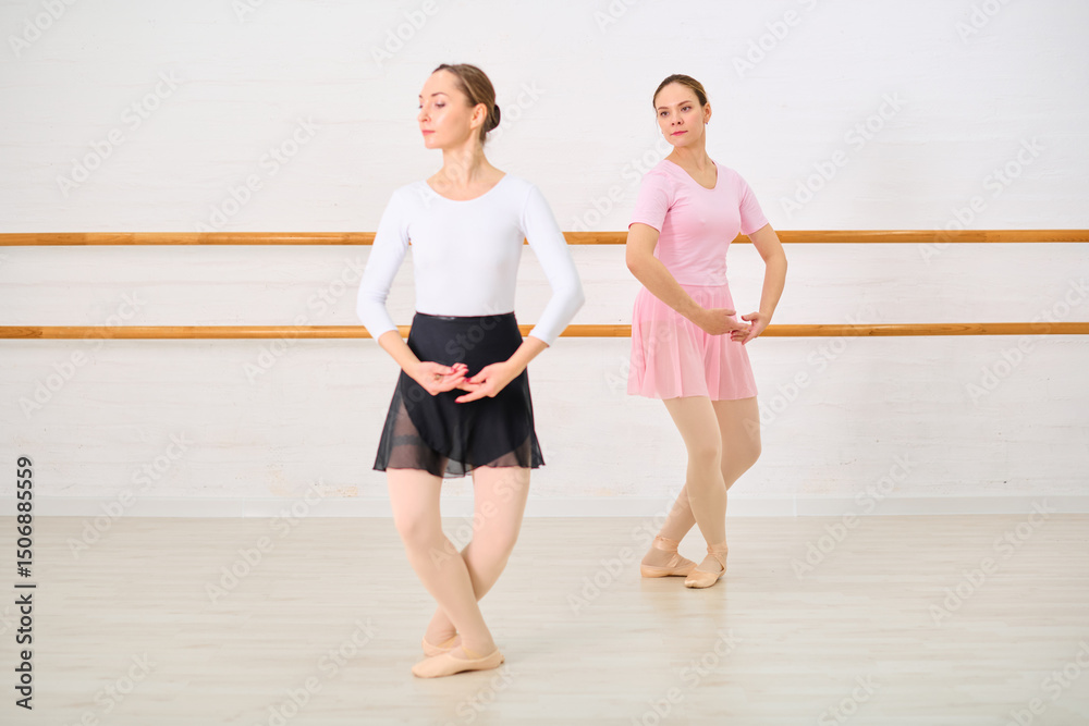Two young female ballerinas with light hair, in graceful poses at ballet barre. One wears white and black, other pink. Bright studio lighting emphasizes their poise Two young female ballerinas with light hair, in graceful poses at ballet barre. One wears white and black, other pink. Bright studio lighting emphasizes their poise