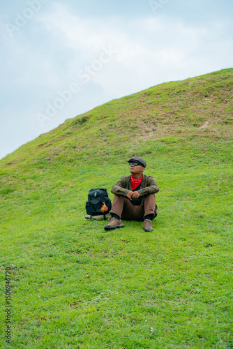 An adventurer is resting by sitting on a stretch of green grass. The Asian man is enjoying the tranquility of nature by resting on a green hill that turns out to be a buried Buddhist temple.