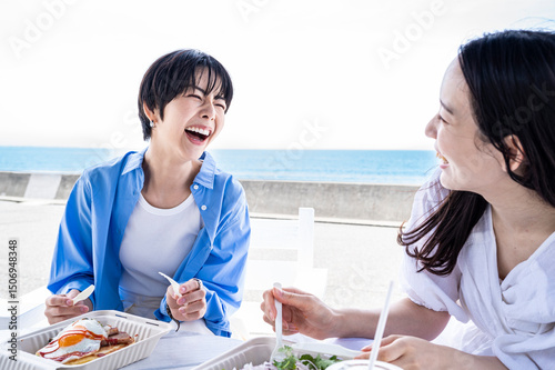 Two Japanese women smiling and laughing while sharing lunch by the ocean, symbolising strong friendship, emotional wellbeing, and healthy lifestyle