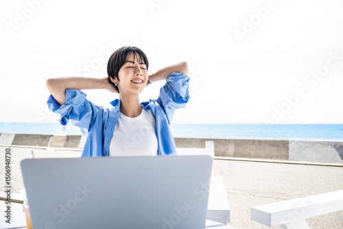 Young Japanese woman working remotely by the seaside using a laptop and smartphone, enjoying a flexible digital nomad lifestyle with technology and ocean views