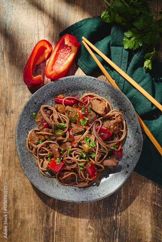 Wok with Soba noodles with turkey, mushrooms and sweet red bell pepper in plate on wooden table in the sun. Asian food, concept of street food. Top view.