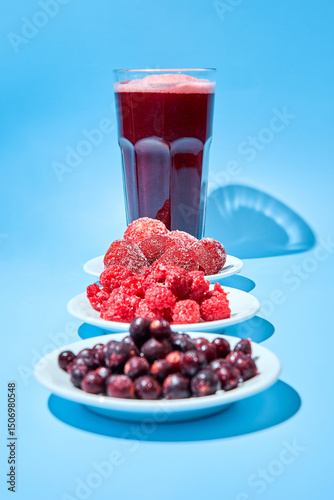 Glass with berry lemonade made from strawberries, frozen raspberries and blackcurrants. Frozen berries lie on small white plates.