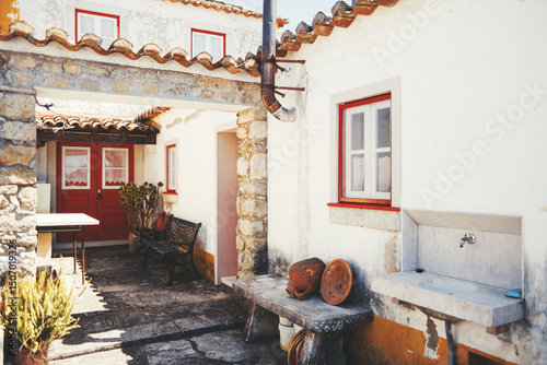 Wallpaper Mural Traditional rustic courtyard in Aldeia da Mata Pequena, Portugal, featuring whitewashed walls, red window frames, stone details, ceramic pots, and an old outdoor sink bathed in sunlight Torontodigital.ca