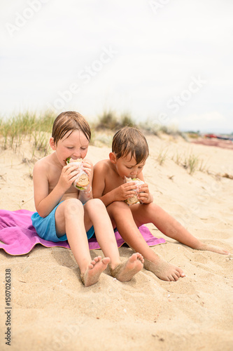 Two wet-haired boys in swim trunks sitting on a towel at the beach, eating sandwiches after swimming. Sunny summer day, childhood moments, seaside snack time.