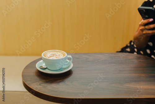 Cup of cappuccino coffee on wooden table with woman using cellphone in background.
