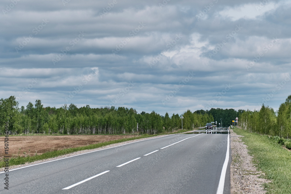 Fototapeta premium Mirage on road going into distance, puddle is seen. Roadside rural landscape. Fata morgana