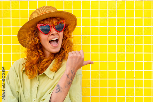 Enthusiastic redhead influencer pointing aside on yellow tiled wall