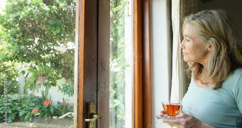 Senior woman enjoying tea by window, gazing thoughtfully at garden, at home