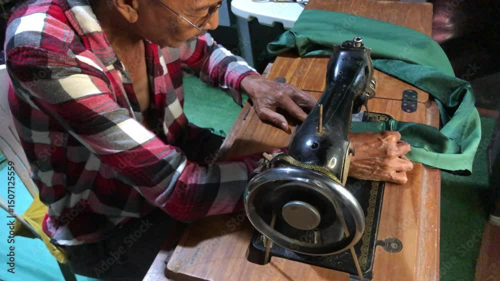 elderly man is sewing a garment with a manually operated, old-fashioned sewing machine