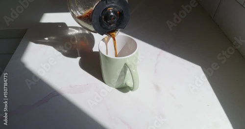 Hot coffee is being poured from a glass carafe into a light green mug sitting on a shiny marble surface, illuminated by warm sunlight from a window.