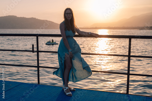 A woman in a flowing green dress poses on a pier by the sea at sunset, with a Turkey flag waving in the background.