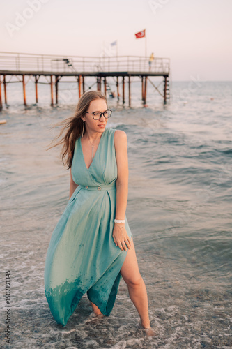 A woman in a flowing green dress poses on a pier by the sea at sunset, with a Turkey flag waving in the background.