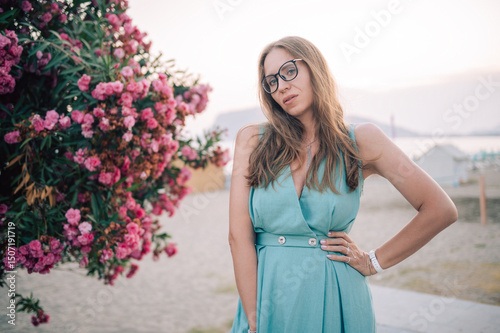 A graceful woman in a turquoise dress poses near blooming pink flowers, enjoying a peaceful moment in a natural outdoor setting.