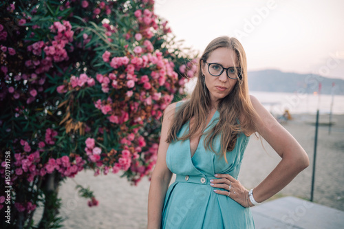 A graceful woman in a turquoise dress poses near blooming pink flowers, enjoying a peaceful moment in a natural outdoor setting.